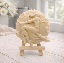 Load image into Gallery viewer, A circular handmade vegan soap featuring a detailed high-relief carving of a witch wearing a pointed hat, flying on a broomstick across a cratered moon, displayed on a miniature wooden easel.