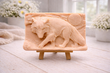 Load image into Gallery viewer, A rectangular handmade vegan soap, featuring a detailed relief carving of a wolf in mid-stride across a rugged landscape with a large full moon in the background, displayed on a miniature wooden easel.