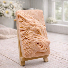 Load image into Gallery viewer, A rectangular handmade vegan soap featuring a highly detailed, 3D relief carving of a wolf's head in profile with a thick, textured mane, displayed on a miniature wooden easel.