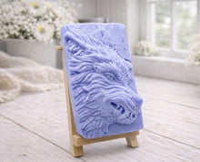 Load image into Gallery viewer, A rectangular handmade vegan soap featuring a highly detailed, 3D relief carving of a wolf's head in profile with a thick, textured mane, displayed on a miniature wooden easel.