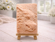Load image into Gallery viewer, A rectangular handmade vegan soap featuring a highly detailed, 3D relief carving of a wolf's head in profile with a thick, textured mane, displayed on a miniature wooden easel.