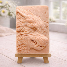 Load image into Gallery viewer, A rectangular handmade vegan soap featuring a highly detailed, 3D relief carving of a wolf's head in profile with a thick, textured mane, displayed on a miniature wooden easel.