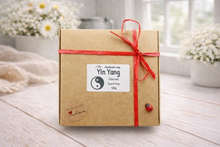 Load image into Gallery viewer, Gift box with a red ribbon and 'Yin Yang' label on a wooden surface with flowers in the background