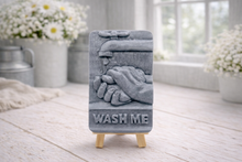 Load image into Gallery viewer, handmade vegan soap featuring a relief sculpture of hands being washed under a dripping tap with the words "WASH ME" at the base, displayed on a small wooden easel in a bright farmhouse decor kitchen setting.
