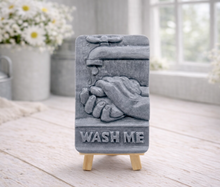 Load image into Gallery viewer, handmade vegan soap featuring a relief sculpture of hands being washed under a dripping tap with the words "WASH ME" at the base, displayed on a small wooden easel in a bright farmhouse decor kitchen setting.