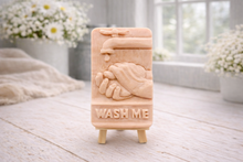 Load image into Gallery viewer, handmade vegan soap featuring a relief sculpture of hands being washed under a dripping tap with the words "WASH ME" at the base, displayed on a small wooden easel in a bright farmhouse decor kitchen setting.