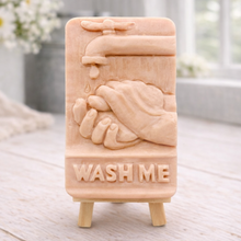 Load image into Gallery viewer, handmade vegan soap featuring a relief sculpture of hands being washed under a dripping tap with the words "WASH ME" at the base, displayed on a small wooden easel in a bright farmhouse decor kitchen setting.