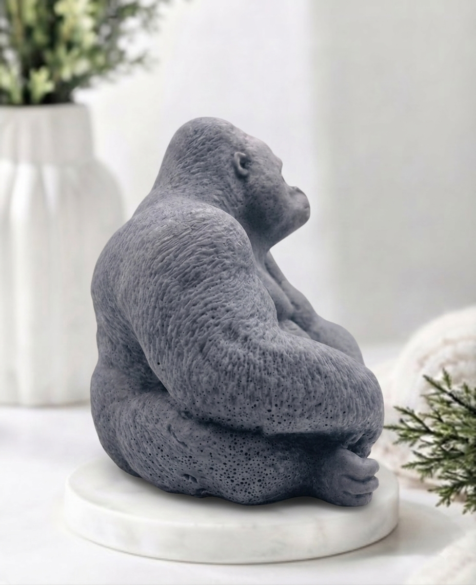 A gray gorilla soap sitting cross-legged on a round white marble base, styled on a light surface with soft-focus plants and decor in the background.