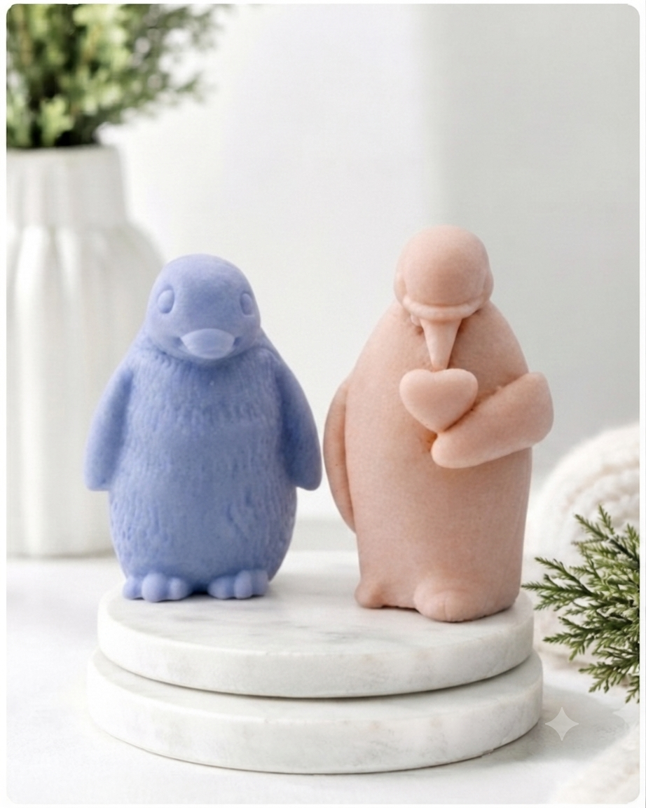 Two cute penguin soaps sitting on a round white marble pedestal. The left penguin is light blue with a textured fuzzy finish, and the right penguin is a soft peach-pink with a smooth matte finish, holding a small heart.
