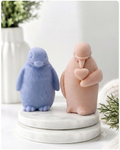 Two cute penguin soaps sitting on a round white marble pedestal. The left penguin is light blue with a textured fuzzy finish, and the right penguin is a soft peach-pink with a smooth matte finish, holding a small heart.
