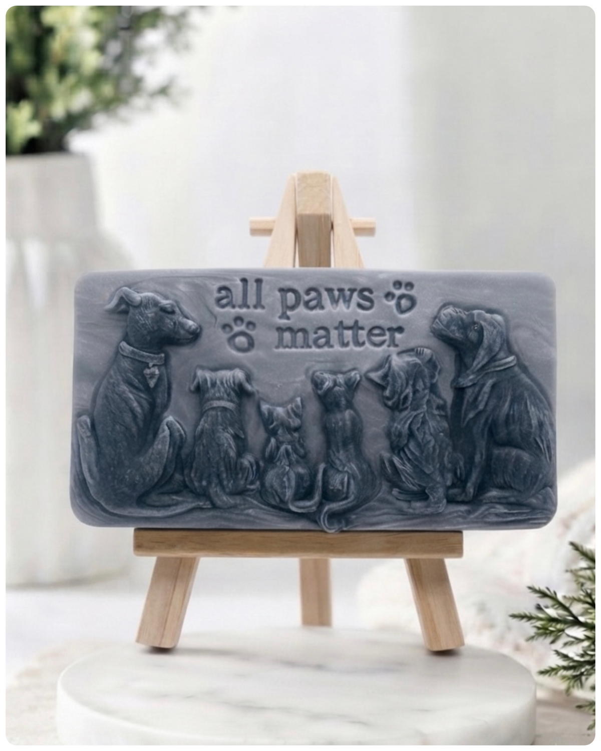 A rectangular handmade vegan soap bar gift, featuring a detailed relief carving of several dogs and a cat sitting together, with the words "all paws matter" and two paw prints embossed at the top, displayed on a wooden easel. Made in Blackpool.