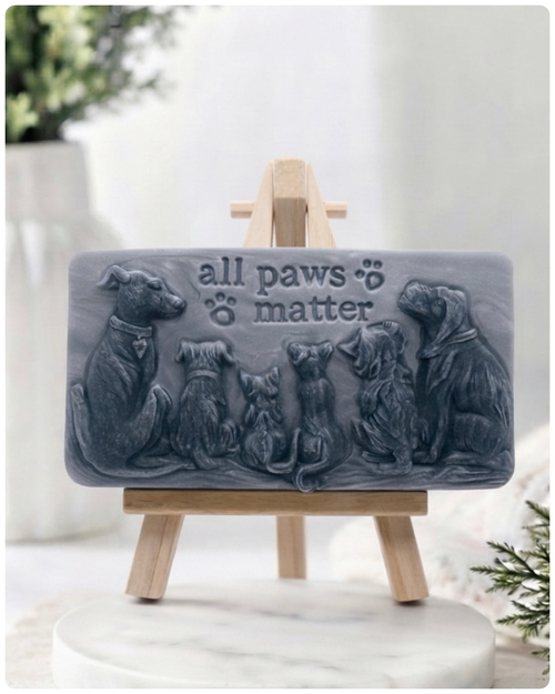 A rectangular handmade vegan soap bar gift, featuring a detailed relief carving of several dogs and a cat sitting together, with the words "all paws matter" and two paw prints embossed at the top, displayed on a wooden easel. Made in Blackpool.