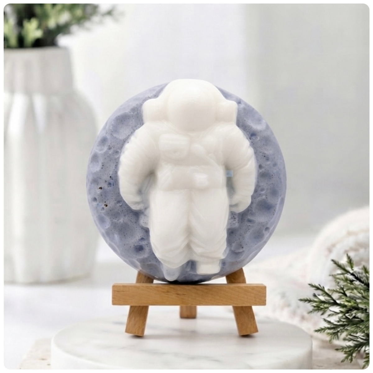 handmade vegan soap in the shape of a realistic astronaut sculpture sitting on the moon, featuring detailed spacesuit textures and craters on the lunar surface, displayed in a bright farmhouse decor setting.