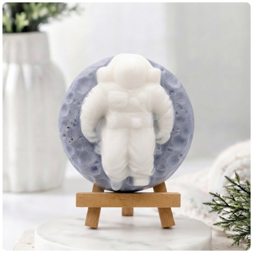 handmade vegan soap in the shape of a realistic astronaut sculpture sitting on the moon, featuring detailed spacesuit textures and craters on the lunar surface, displayed in a bright farmhouse decor setting.