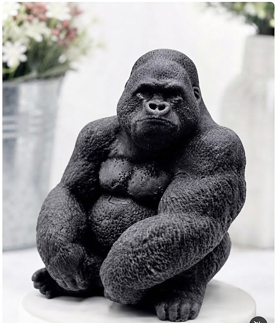A large black gorilla vegan soap gift sitting on a white pedestal, set against a softly blurred indoor background with potted plants.