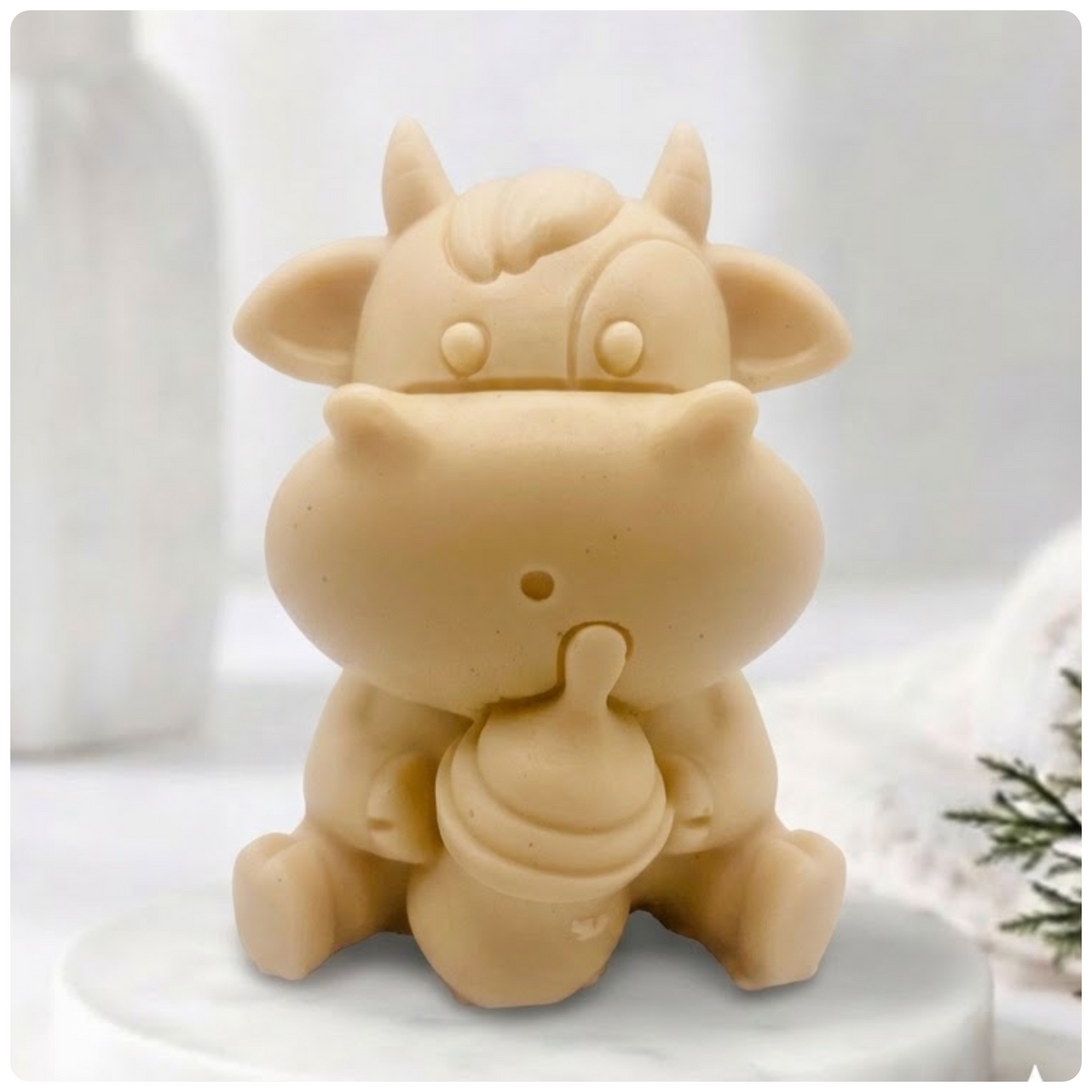 A baby cow-shaped soap holding a small milk bottle, photographed on a soft white background with a blurred bottle and greenery in the surroundings.