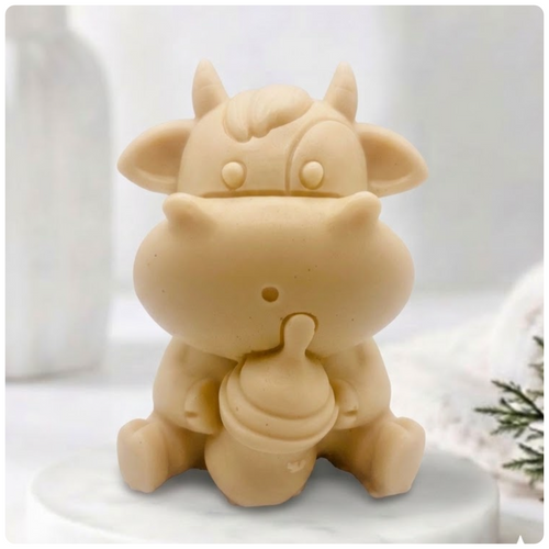 A baby cow-shaped soap holding a small milk bottle, photographed on a soft white background with a blurred bottle and greenery in the surroundings.