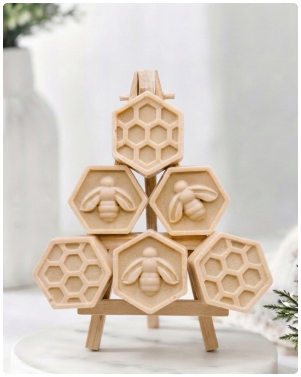 A handmade vegan soap gift set featuring six hexagonal bars stacked in a pyramid on a small wooden easel; some bars show a honeybee relief and others a honeycomb pattern