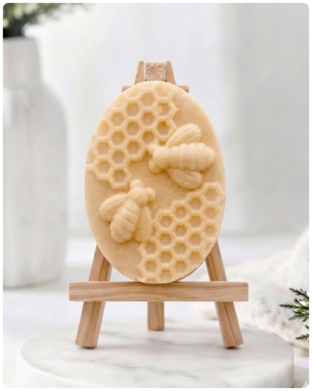handmade vegan soap featuring an oval relief sculpture of two honey bees crawling on a honeycomb pattern, displayed on a small wooden easel in a bright farmhouse decor setting.