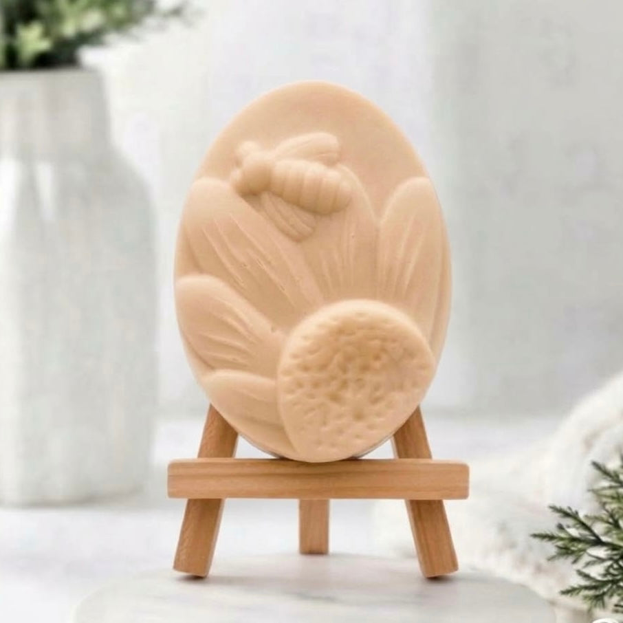 A yellow oval handmade soap bar with a raised bee and leaf design, displayed on a small wooden stand against a soft, blurred white background.