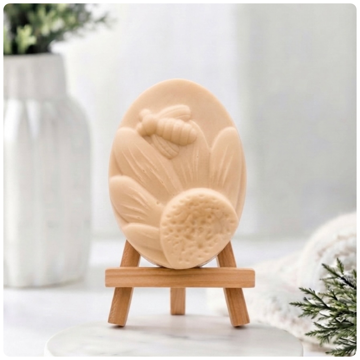 A handmade vegan soap in a natural colour, featuring an oval relief of a honeybee landing on a large sunflower with detailed petals and a textured centre, displayed on a small wooden easel.