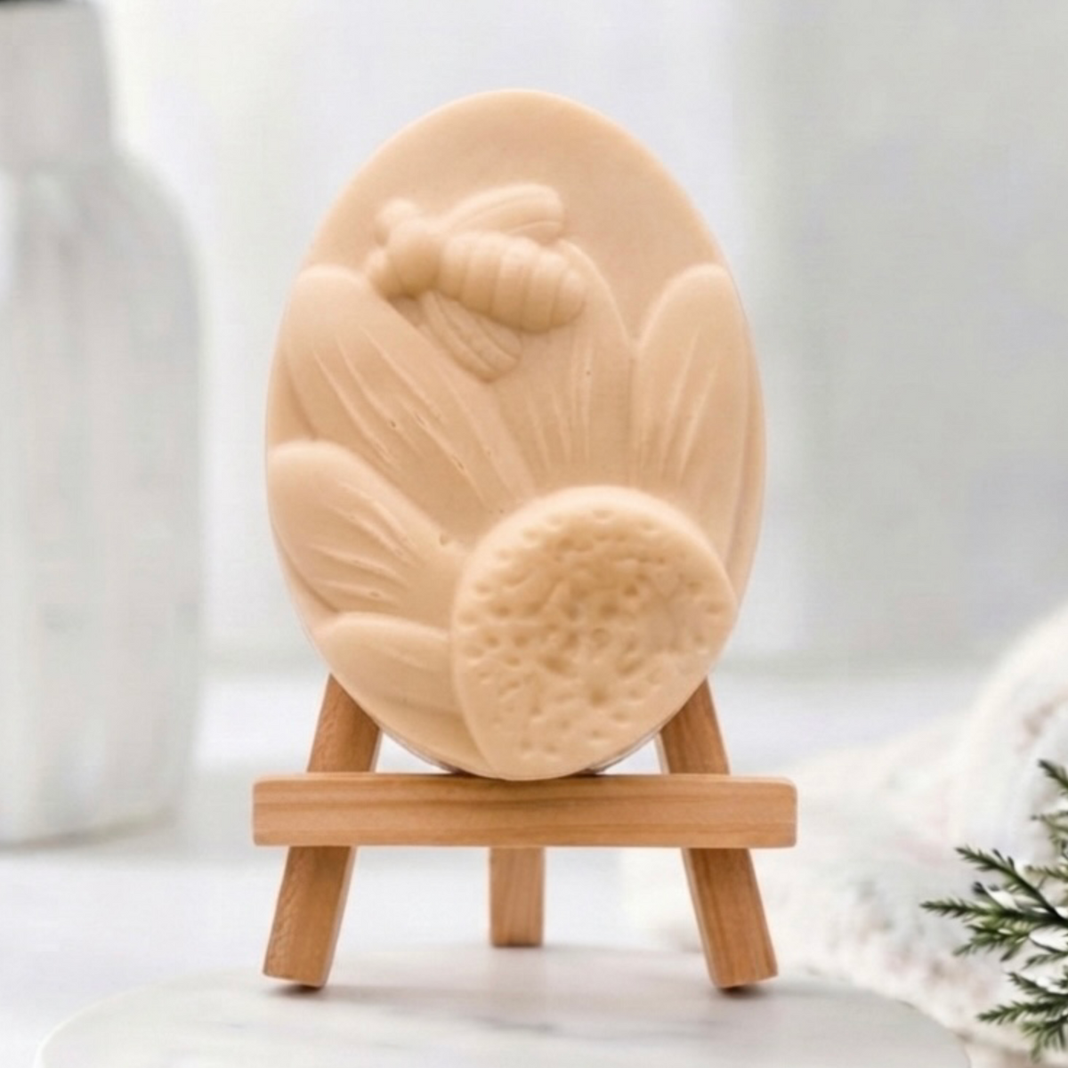 A handmade vegan soap in a natural colour, featuring an oval relief of a honeybee landing on a large sunflower with detailed petals and a textured centre, displayed on a small wooden easel.