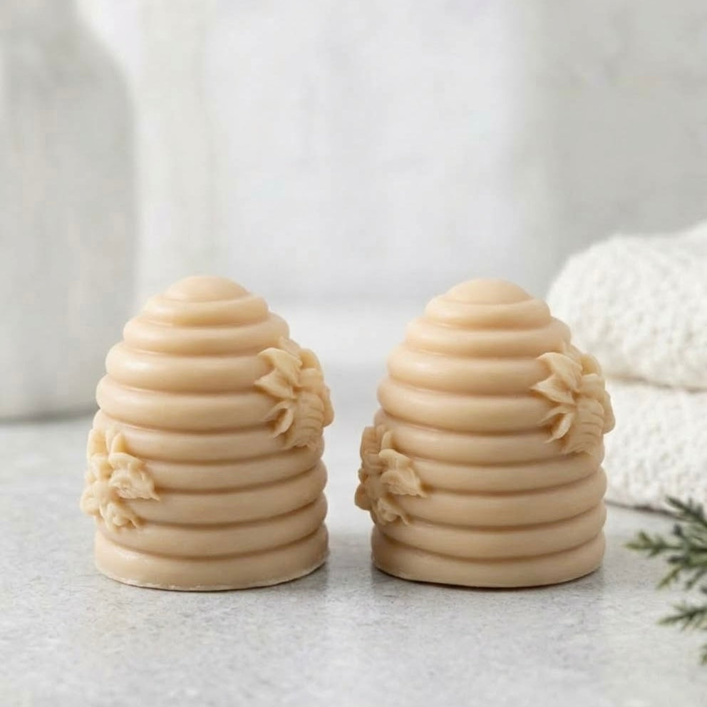 Two soft yellow coloured soaps shaped like beehives, shown side by side on a light grey surface with a soft, blurred background.