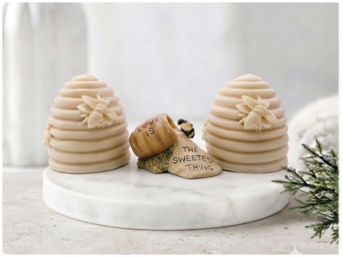 Two handmade vegan soaps sculpted into traditional dome-shaped beehives (skeps) with small bees resting on the sides, flanking a decorative centrepiece featuring a honey pot and the words "The Sweetest Thing," all displayed on a rustic wooden surface.