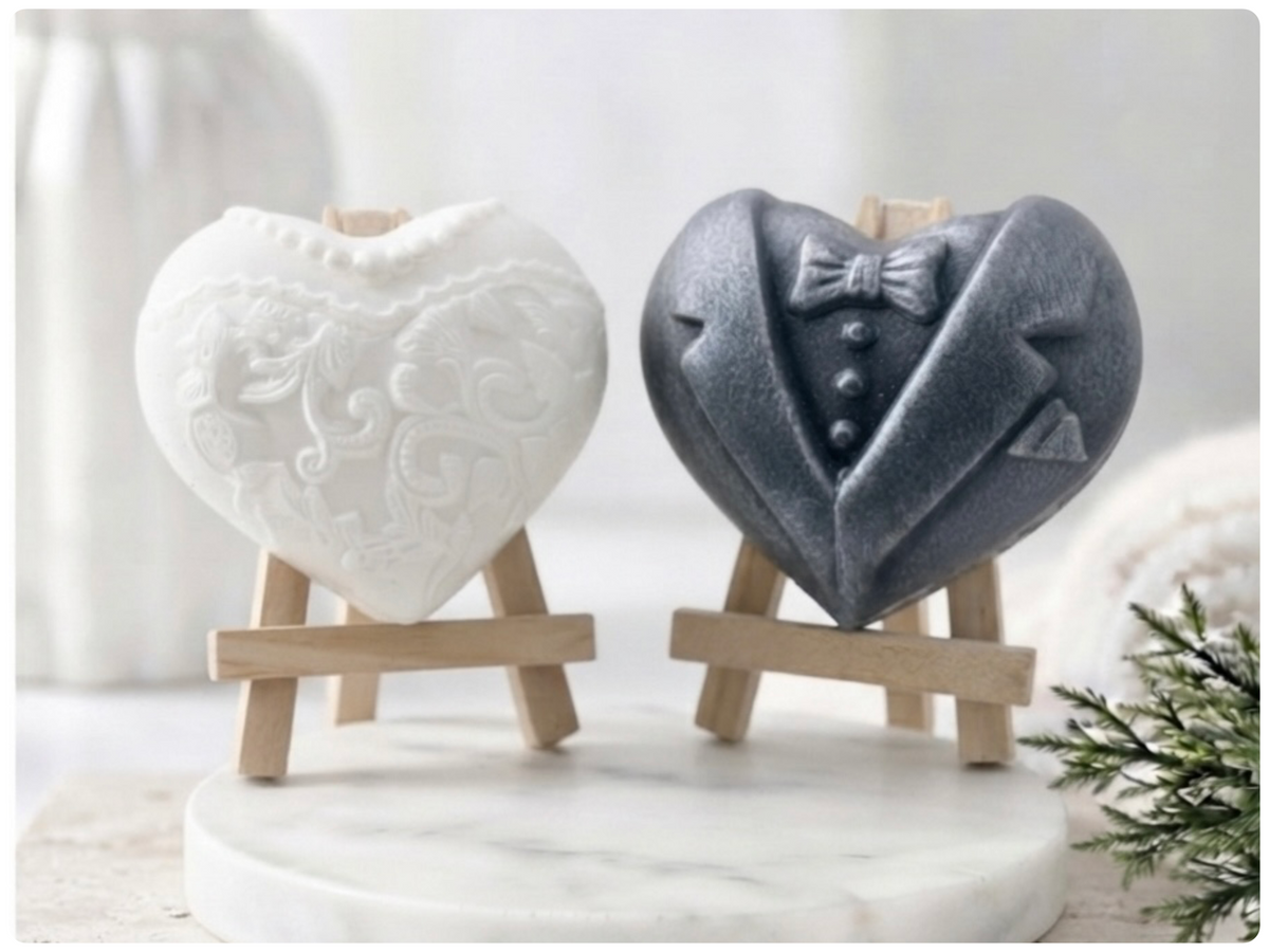 A pair of heart-shaped wedding soaps shaped like a bride's white lace dress and a groom's grey tuxedo, displayed on small wooden easels. Gift boxed ready to give. 