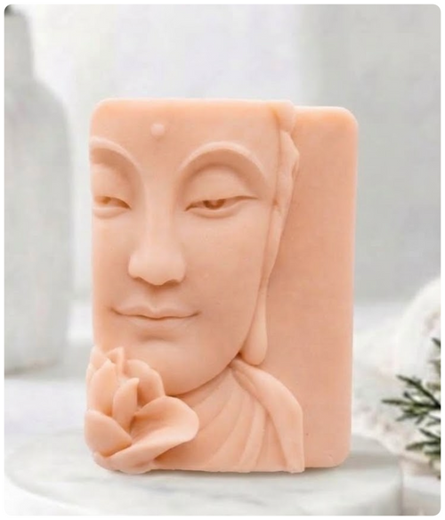A detailed carved pastel pink French clay soap bar depicting the half-face of a serene Buddha holding a lotus flower near the chin. It sits on a round marble pedestal against a soft white interior background.