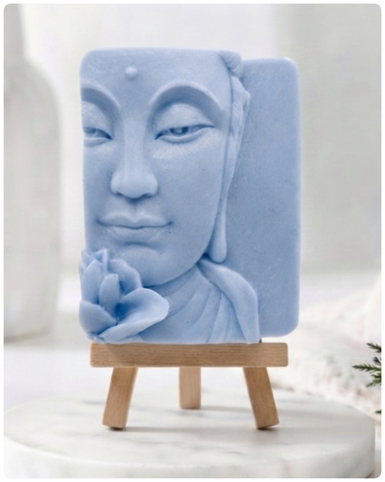 handmade vegan soap featuring a serene Buddha face relief with a lotus flower, displayed on a small wooden easel in a bright farmhouse decor setting with flowers and a rustic milk churn.