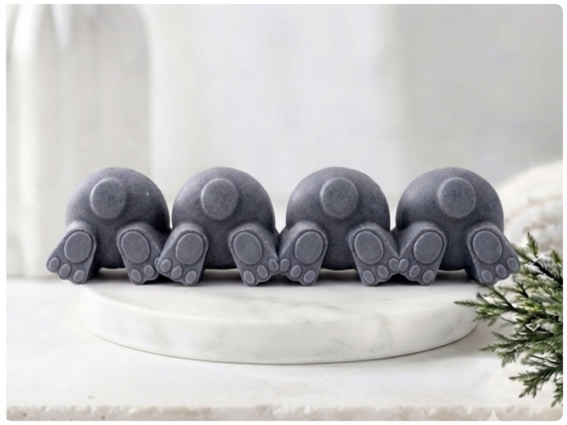 A set of four handmade vegan soaps in the shape of fluffy bunny tails and paws, displayed in a row on a neutral linen cloth with a basket of Easter eggs in the background.