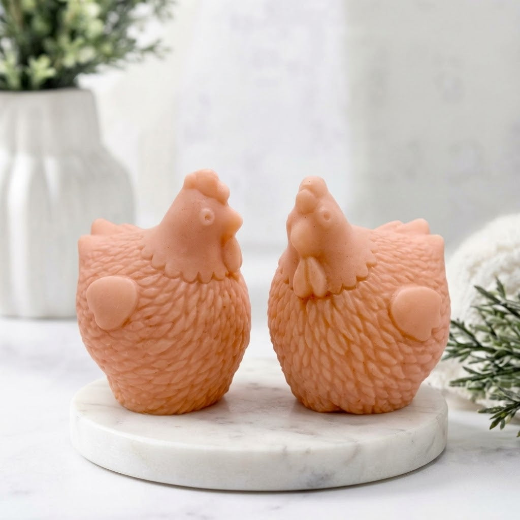 A close-up product photo of a pair of detailed chicken-shaped soap bars from the "Little Chickens" gift set, placed on a white marble display stand with a blur background of a flower vase.