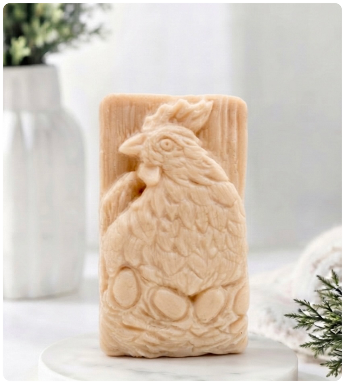 A rectangular handmade vegan soap in a natural colour, featuring a highly detailed relief carving of a broody hen chicken sitting on a nest of eggs, displayed upright on a light wooden surface.