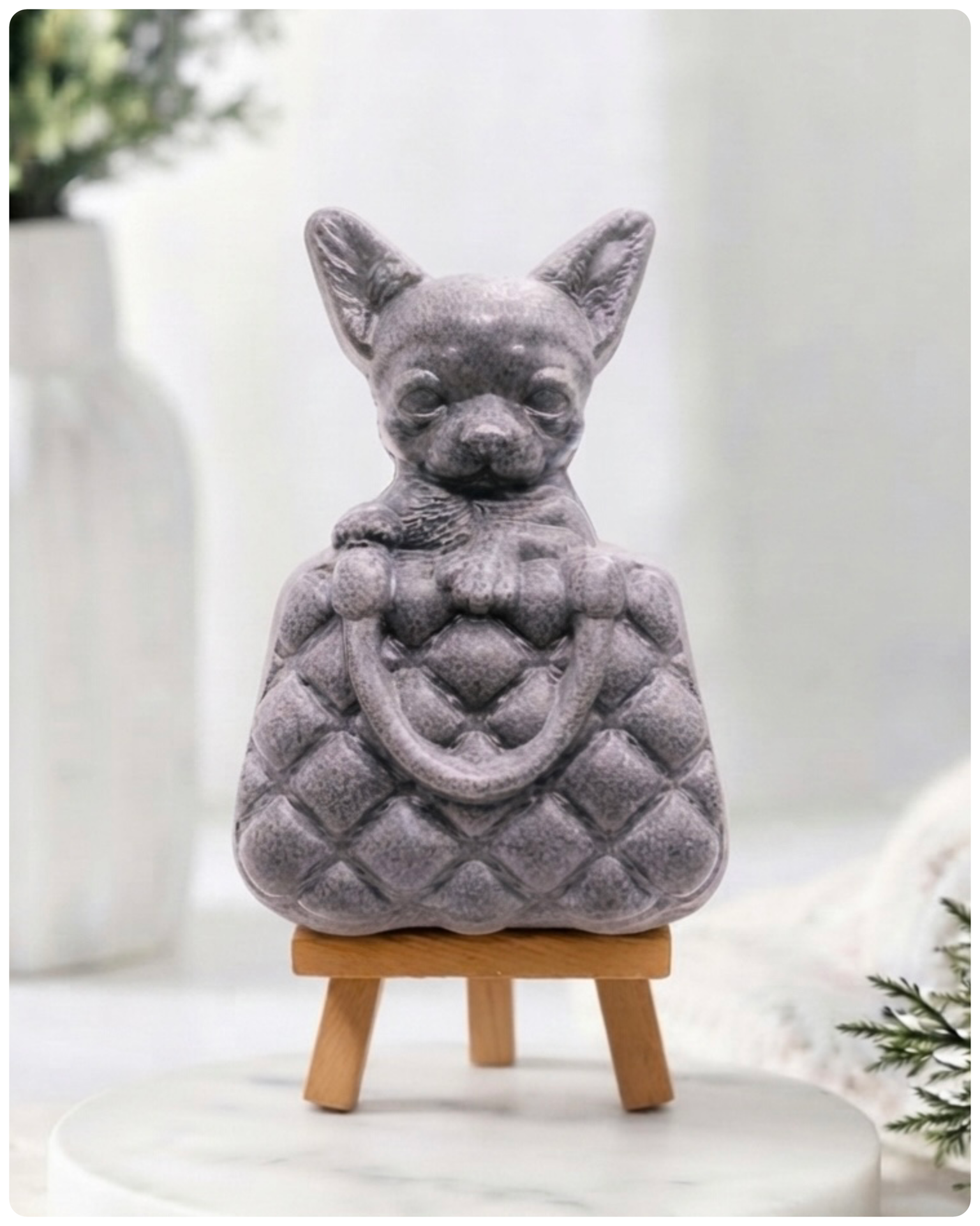 A highly detailed handmade vegan soap in a speckled grey marble finish, sculpted as a small chihuahua puppy with large ears peeking out from inside a quilted designer-style handbag, displayed on a miniature wooden easel.