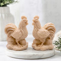 A detailed shot of two light yellow-colored cockerel soaps, showcasing their intricate, natural texture and the fine feather detailing of the wattle, comb, and large tail feathers as they stand on a white marble surface.