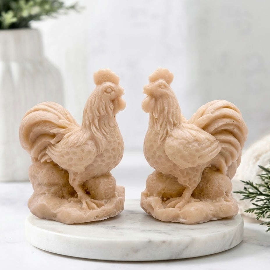 A detailed shot of two light yellow-colored cockerel soaps, showcasing their intricate, natural texture and the fine feather detailing of the wattle, comb, and large tail feathers as they stand on a white marble surface.