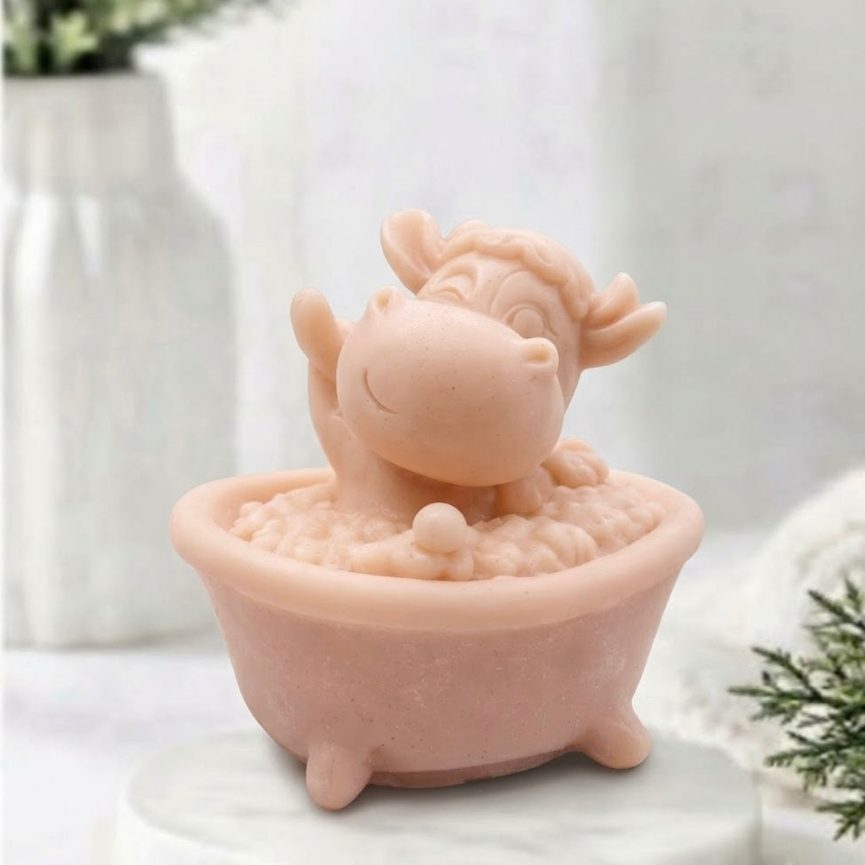 Novelty vegan soap bar moulded into a cute pale pink cartoon cow waving happily while sitting in a miniature footed bathtub surrounded by bubbles.