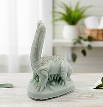 Load image into Gallery viewer, diplodocus dinosaur vegan soap figurine on a wooden surface with a blurred indoor background.
