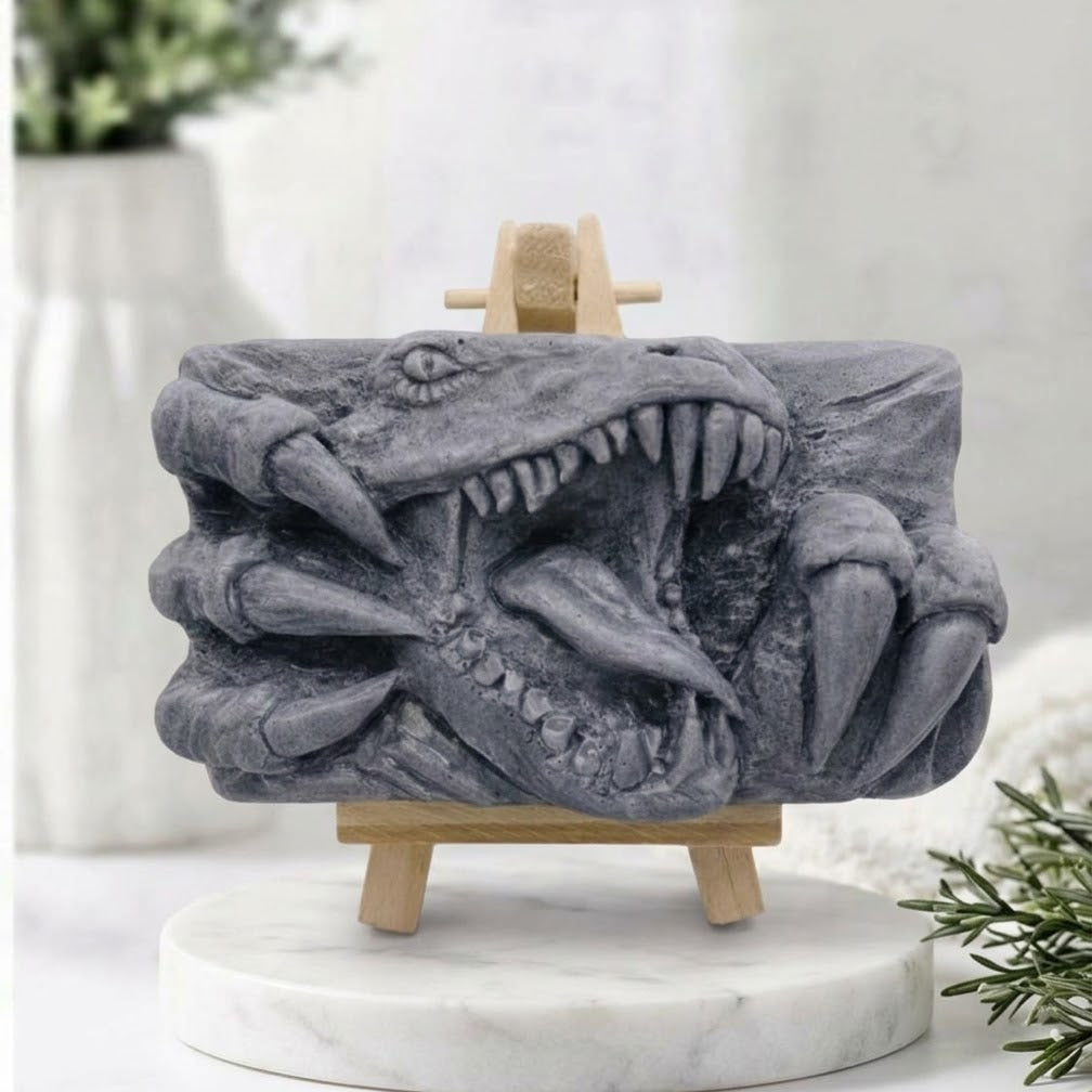 A charcoal grey rectangular handmade vegan soap  featuring a high-relief carving of a Tyrannosaurus Rex with its jaws wide open in a roar and sharp claws framing the face, displayed on a miniature wooden easel.
