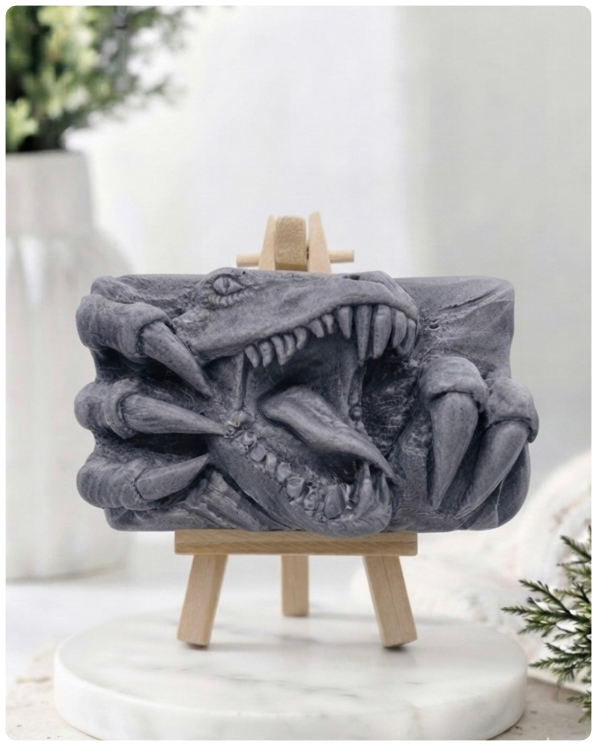 A rectangular handmade vegan soap  featuring a high-relief carving of a Tyrannosaurus Rex with its jaws wide open in a roar and sharp claws framing the face, displayed on a miniature wooden easel.