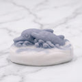An up-close view of an oval, artisan vegan soap bar featuring two gray-blue dolphins swimming over white waves, resting on a white marble surface.