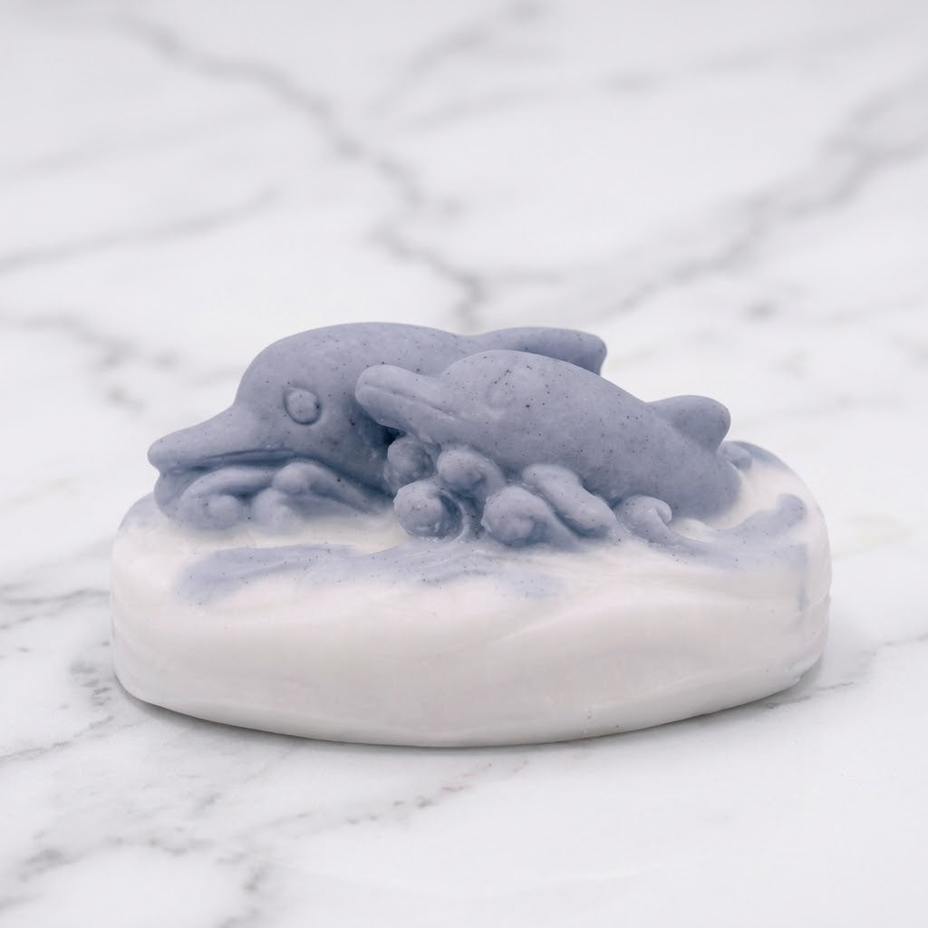 An up-close view of an oval, artisan vegan soap bar featuring two gray-blue dolphins swimming over white waves, resting on a white marble surface.