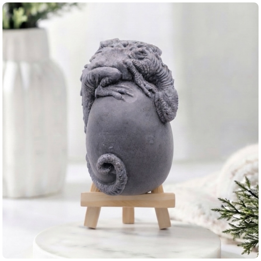 A 3D handmade vegan soap in a charcoal-grey tone, sculpted as a large prehistoric egg with a small dinosaur or dragon clinging to the top, featuring detailed scales and a curled tail, displayed on a miniature wooden easel.
