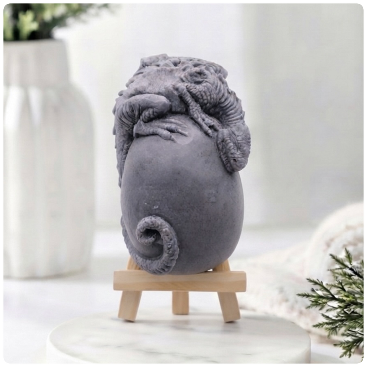 A 3D handmade vegan soap in a charcoal-grey tone, sculpted as a large prehistoric egg with a small dinosaur or dragon clinging to the top, featuring detailed scales and a curled tail, displayed on a miniature wooden easel.