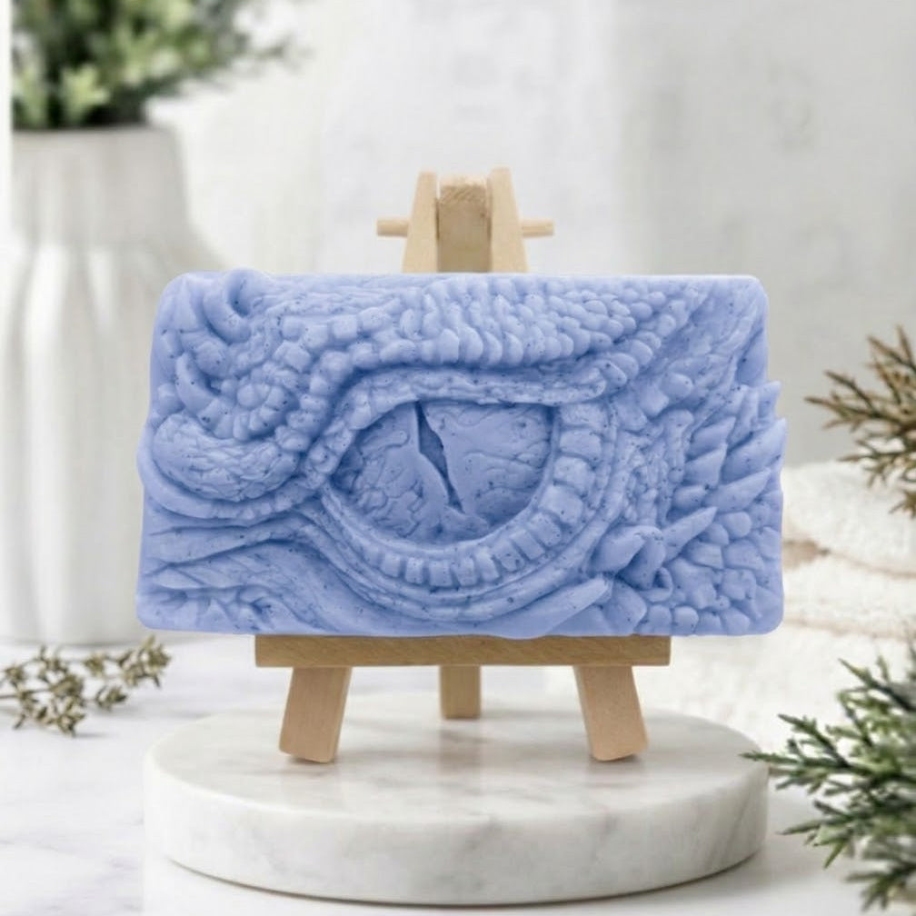 A rectangular handmade vegan soap in a soft blue tone, featuring a highly detailed, close-up relief of a mythical dragon or dinosaur eye surrounded by thick, pebbled scales and reptilian skin textures, displayed on a miniature wooden easel.