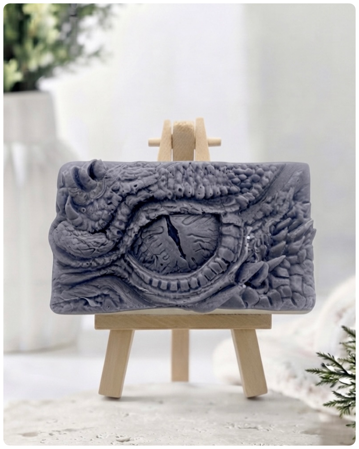 A rectangular handmade vegan soap in a slate-grey tone, featuring a highly detailed, close-up relief of a mythical dragon or dinosaur eye surrounded by thick, pebbled scales and reptilian skin textures, displayed on a miniature wooden easel.