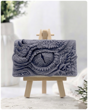 Load image into Gallery viewer, A rectangular handmade vegan soap in a slate-grey tone, featuring a highly detailed, close-up relief of a mythical dragon or dinosaur eye surrounded by thick, pebbled scales and reptilian skin textures, displayed on a miniature wooden easel.
