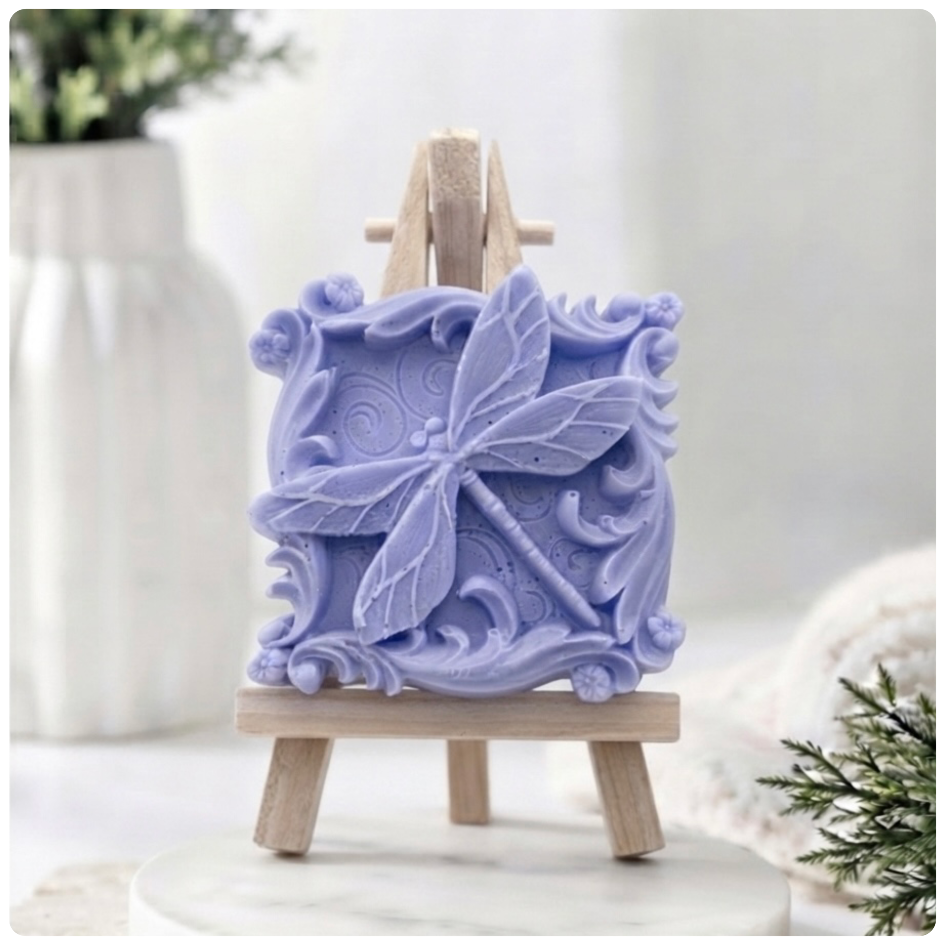 A beautiful handmade vegan soap in a natural  tone, featuring a highly detailed relief of a dragonfly with delicate, outspread wings and a textured body, displayed on a miniature wooden easel against a soft farmhouse background.