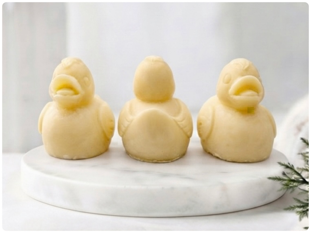 Three handmade vegan soaps in the shape of fluffy ducklings with open beaks, displayed on a neutral linen cloth in a bright farmhouse decor setting with Easter eggs in the background.